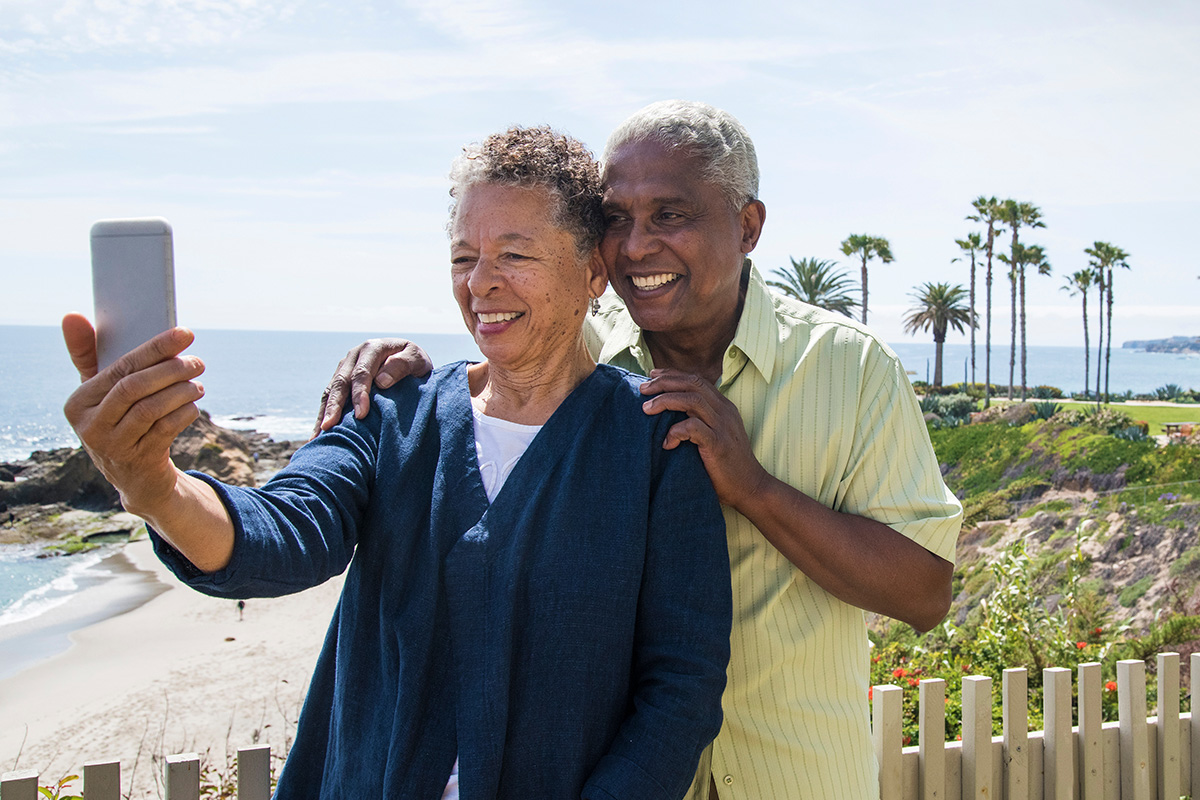 Smiling senior couple taking a selfie together at a scenic beachside location with palm trees and ocean in the background.