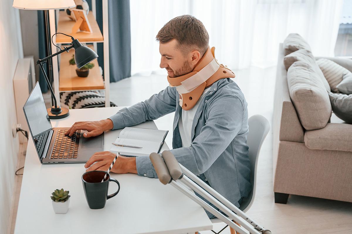 Injured man with a neck brace using a laptop at home, with crutches beside him and a notebook on the desk.