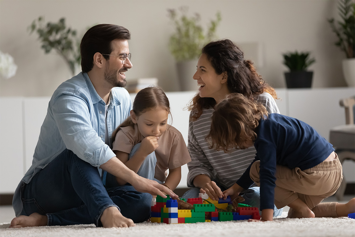 Happy parents sitting on the floor playing with two young children and colorful building blocks in a cozy living room.