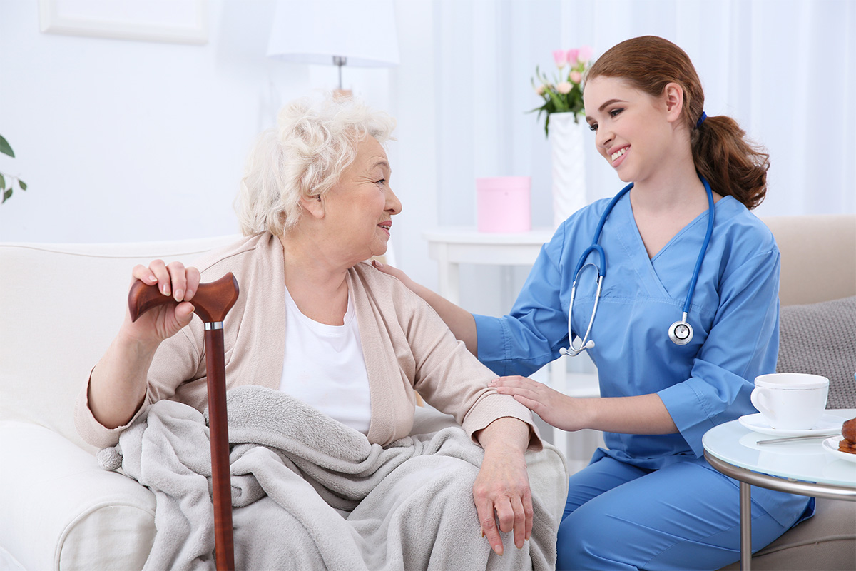 Young nurse in scrubs gently touching the arm of a smiling elderly woman with a cane, sitting together in a bright living room.