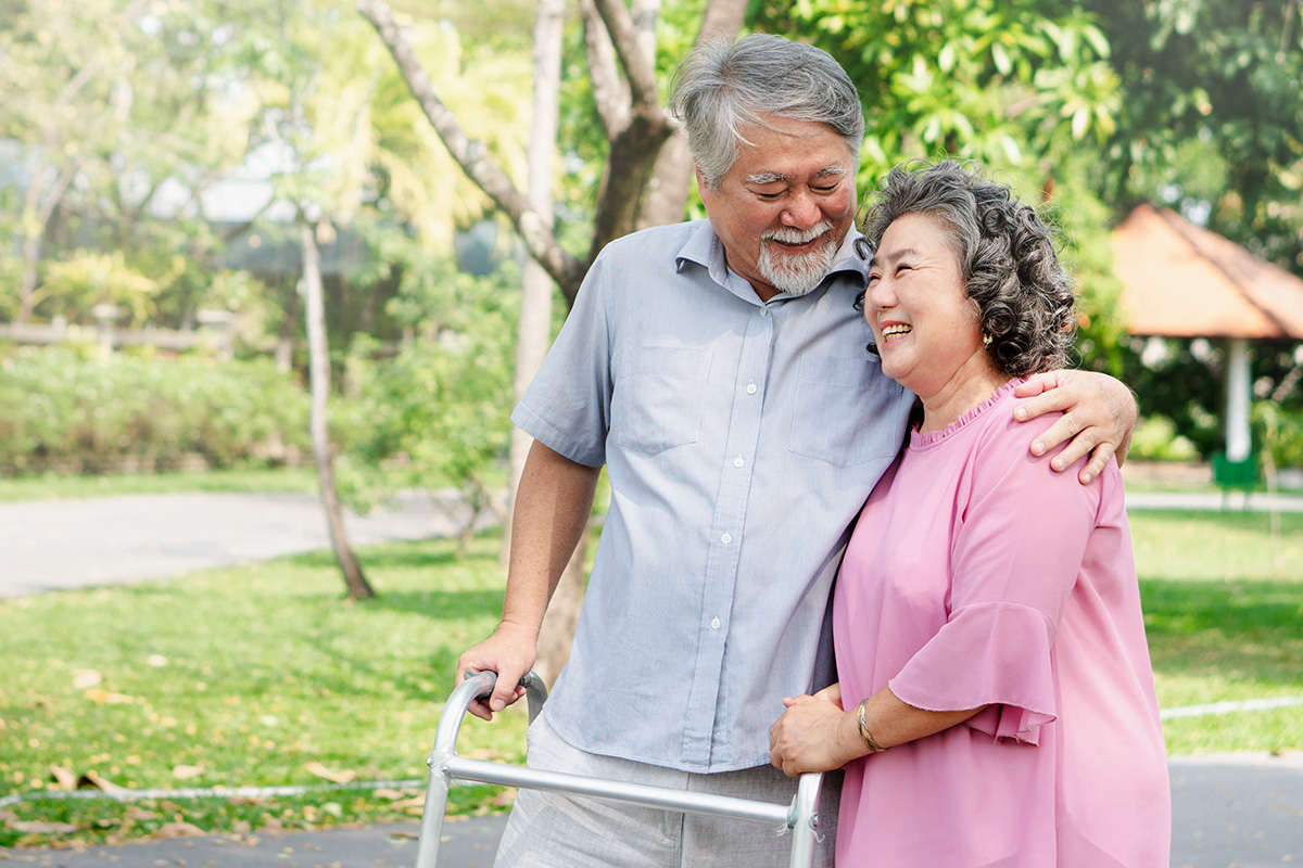 Elderly couple smiling and walking together in a park, with one using a walker and the other embracing them.
